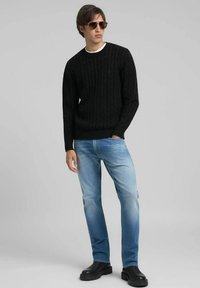 Pull noir en maille torsadée, jean bleu clair et bottes épaisses noires. Le mannequin porte des lunettes de soleil marron. Fond simple.