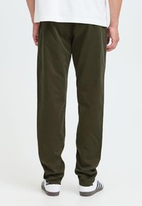 Homme portant un pantalon vert foncé, une chemise blanche et des baskets blanches avec des rayures noires, debout de dos sur un fond uni.