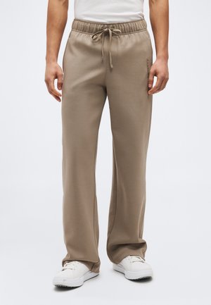 Homme portant un pantalon de survêtement beige à cordon de serrage et des baskets blanches, debout devant un fond clair uni.