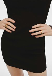 Anillos de oro con diseños intrincados llevados en los dedos, sobre un vestido negro liso. Superficie suave; visibles diversas formas y estilos.