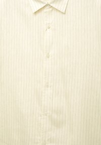 Chemise légère à rayures fines en tissu doux. Présente un boutonnière à l'avant et un col rabattu. La couleur est écrue avec de fines rayures beiges.