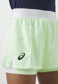 Ljusgröna sportshorts med en vit midja, utrustade med en svart logotyp på vänster sida och en lager-design för komfort.