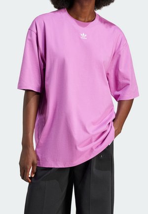 Osoba nosząca luźny, lawendowy t-shirt Adidas z małym białym logo na piersi, w czarnych spodniach, stojąca z jedną ręką w kieszeni.