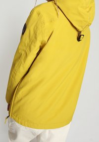 Gele waterdichte jas met aantrekkoordkap, zijrits en gestructureerde stof. Voorzien van een ronde logopatch op de mouw.