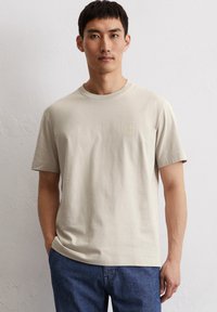 Beige Baumwoll-T-Shirt mit kurzen Ärmeln und Rundhalsausschnitt. Mit einem dezenten, geprägten Logo auf der Brust. Weiche Textur und bequeme Passform.