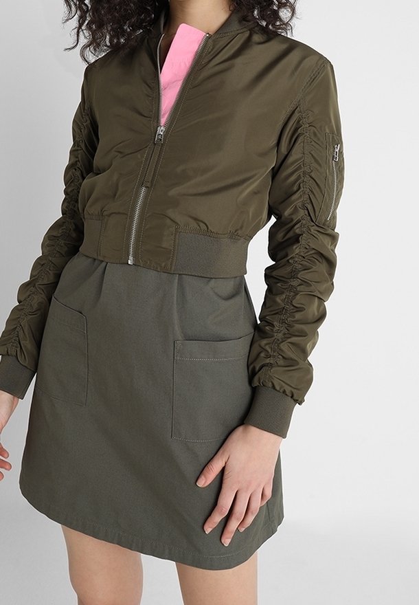 Kobieta ubrana w oliwkową kurtkę bombkę z krótkim rękawem, na której ma dopasowaną oliwkową spódnicę z przednimi kieszeniami i widocznym różowym kołnierzykiem koszuli.