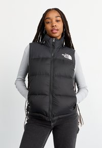 Vest negru căptușit cu guler înalt, fermoar frontal și logo pe pieptul stâng. Purtați peste un top gri cu mânecă lungă, afișat din față.