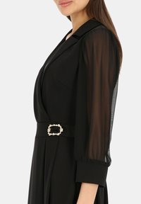 Czarna sukienka z przezroczystymi, rękawami do łokcia, dekoltem w kształcie litery V oraz szerokim paskiem z ozdobną, kwadratową klamrą z dodatkami.
