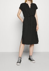 Robe en lin noire à manches courtes, décolleté en V, taille nouée, fentes latérales, portée avec des baskets montantes noires. Texture lisse, design décontracté.