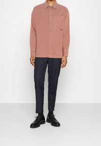 Koszula zapinana na guziki w kolorze rdzy z przednią kieszenią, zestawiona z ciemnymi spodniami w prążki i czarnymi butami. Gładka tkanina, klasyczny kołnierz.