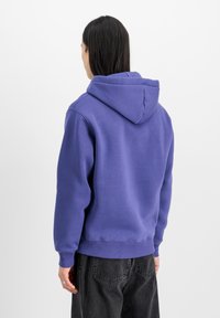 Lila huvtröja med kängurufickor, dragsko på huvan, ribbade cuffs och nederkant. Tyget är mjukt, av bomullsblandning, och har en slät yta.