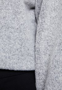 Maglione lavorato a maglia grigio chiaro con una finitura texture, caratterizzato da un scollo rilassato e un tessuto morbido che scivola. Tessuto nero visibile sotto.