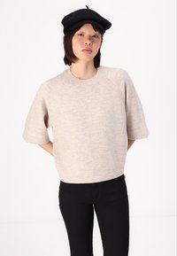 Pulover tricotat bej cu mânecă scurtă, guler rotund și finisaj texturat, asortat cu pantaloni negri. Croială simplă, pătrată, cu o potrivire lejeră.