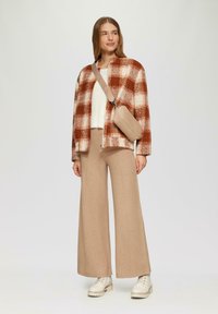 Karierte Überbluse in Rost und Weiß, cremefarbener Strickpullover, beiger weit geschnittener Hose und hellen Schuhen; trägt eine taubfarbene Umhängetasche.