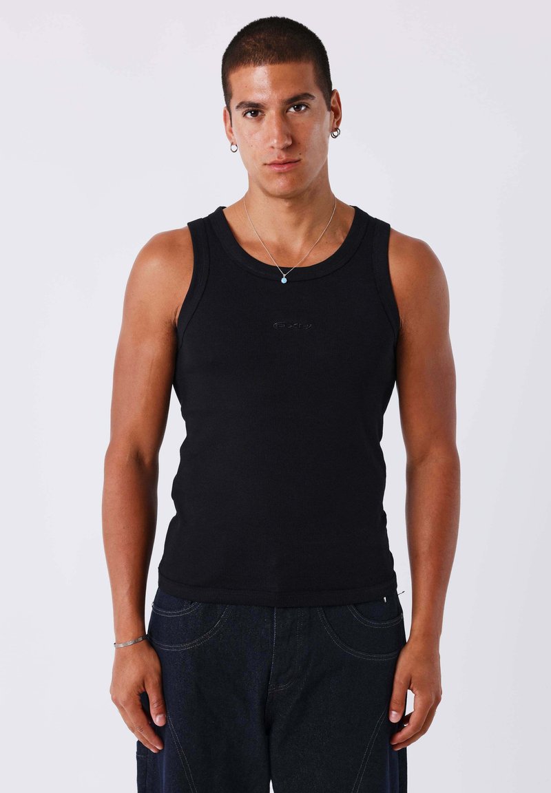 Jeune homme aux cheveux courts portant un haut noir sans manches, un jean foncé, un collier en argent et des boucles d'oreilles en anneau, debout devant un fond uni.