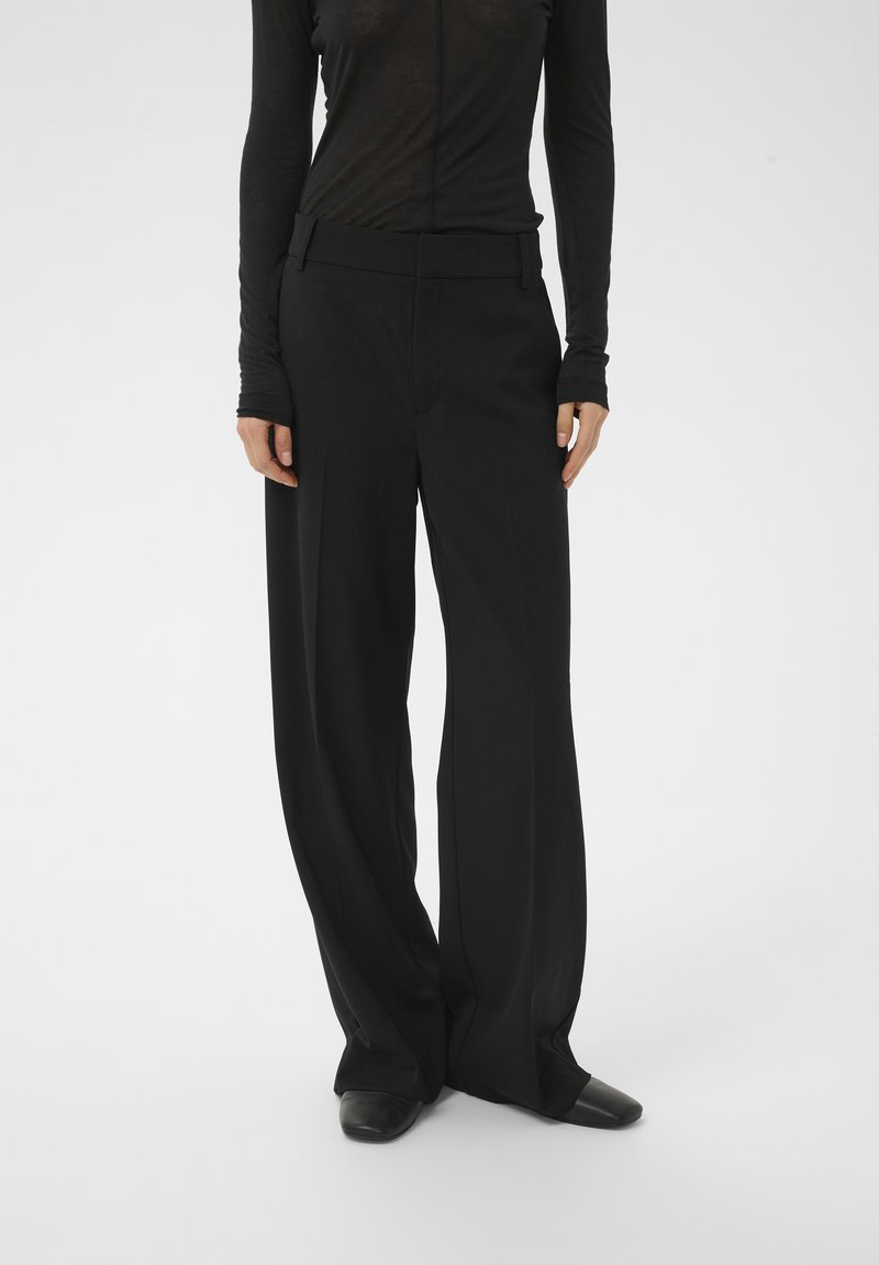 Schwarze, weit geschnittene Hose aus glattem Stoff mit tailliertem Schnitt, flacher Vorderseite und schlankem Bund, kombiniert mit schwarzen Schuhen.