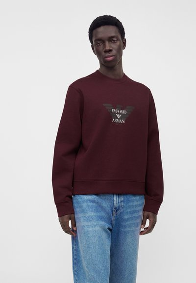 Άνδρας που φοράει μπορντό φούτερ Emporio Armani και ανοιχτό μπλε τζιν, στέκεται μπροστά σε απλό γκρι φόντο.