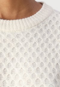 Cremfarbener gestrickter Pullover mit einem strukturierten Wabenmuster und einem gerippten Ausschnitt. Der Stoff wirkt weich und leicht.