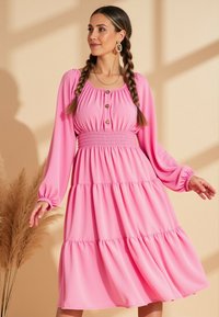 Różowa sukienka z długimi rękawami i marszczonym wpasowaniem w talii. Posiada detale w postaci guzików i falbaniaste spódnice. Wykonana z lekkiego, zwiewnego materiału.