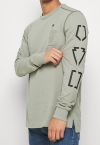 Langärmliges Hemd aus gedämpftem grünem Baumwollstoff, mit schwarzen Grafiken auf den Ärmeln und einem kleinen Logo auf der Brust. Nahtverarbeitung mit Zierstichen.