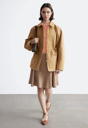 Mujer vestida con una chaqueta color beige sobre un polo coral, falda topo, zapatos marrones con tiras, sosteniendo un pequeño bolso de hombro marrón oscuro, caminando hacia la cámara.