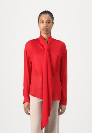 Red silk blouse with a tied neck detail, long sleeves, and a slightly sheer texture, paired with beige trousers.