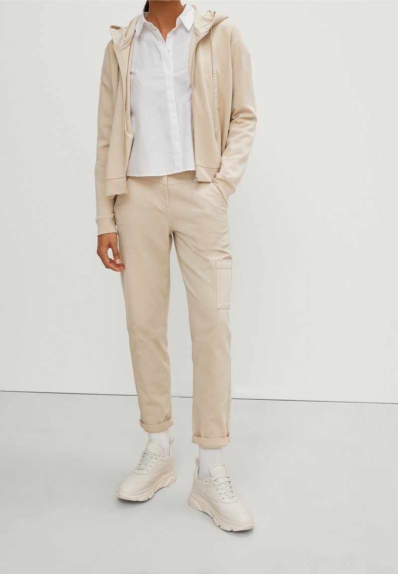 Personne portant un pantalon beige à ourlets retroussés, un sweat à capuche beige zippé, un t-shirt blanc, des chaussettes blanches et des baskets blanches épaisses sur un fond uni.