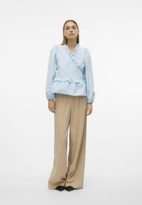Blusa avvolgente azzurro chiaro con dettagli a volant, maniche a sbuffo e vita a peplo, abbinata a pantaloni a gamba larga beige e scarpe nere.