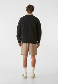 Melna sporta krekls ar ribotām piedurknēm, brūnu rūtainu šortu, krēma zeķes un bēša apavi, rādīts no aizmugures uz vienkārša fona.