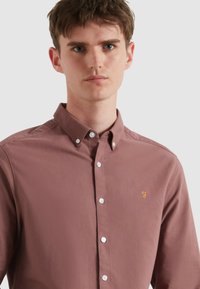 Langsleeve, knoopshirt in stoffige rozenkleur, gemaakt van gladde stof. Beschikt over een klein geel logo en witte knopen aan de voorkant.