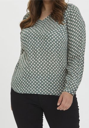 Frau mit grüner Langarmbluse mit kleinem weißem Blumenmuster und schwarzer Hose, von den Schultern bis zu den Hüften gezeigt.