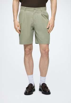 Homme portant un short vert clair sur mesure, des chaussettes blanches hautes et des mocassins en cuir marron foncé à pompons, debout devant un fond blanc.