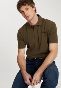 Oliwkowa koszulka polo wykonana z bawełny, z kołnierzykiem, trzema guzikami i haftowanym logo na piersi. Noszona z niebieskimi dżinsami.