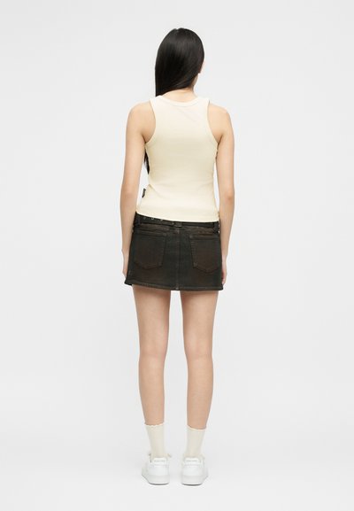 Γυναίκα με μακριά σκούρα μαλλιά που φοράει αμάνικη μπεζ μπλούζα, μαύρη τζιν μίνι φούστα, λευκές κάλτσες και λευκά αθλητικά παπούτσια, στέκεται με την πλάτη προς τα εμπρός.