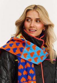 Pletená šatka v jasných farbách s prestriedanými vzormi srdiečok v oranžovej, modrej a akvamarínovej farbe, prehodená cez čiernu prešívanú bundu s červenými akcentmi.