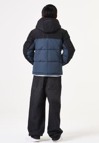 Pufferjack met een blauwe body en zwarte schouders, voorzien van een capuchon en een doorgestikte ontwerp. Gecombineerd met losse, donkere denim broeken en witte sneakers.