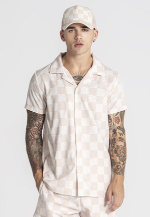 Ung mand med ærmetatoveringer iført beige og hvid ternet kortærmet skjorte, matchende shorts og kasket, stående foran en ensartet baggrund.