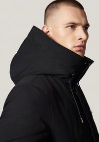 Czarna kurtka z kapturem i wysokim kołnierzem, o gładkiej fakturze i zamknięciu na zamek z przełącznikami. Minimalistyczny design i smukła sylwetka.
