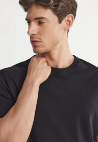 Svart kortärmad t-shirt i slät väv, med klassisk rund halsringning och subtila sömnadsdetaljer, som bärs av en person.