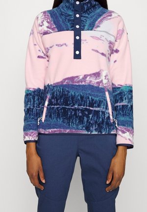 Person trägt einen pinken, lila und blauen Fleece-Pullover mit halbgeknöpfter Vorderseite und Reißverschlusstaschen, kombiniert mit dunkelblauer Hose.