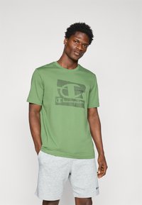 Grünes Kurzarm-T-Shirt aus Baumwolle mit einem strukturierten schwarzen Champion-Logo-Design. Kombiniert mit grauen Sportshorts mit gesticktem Logo.