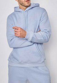 Haut à capuche bleu clair en tissu doux, avec une poche avant, un capuchon à cordon de serrage et des poignets et un ourlet côtelés. Texture lisse.