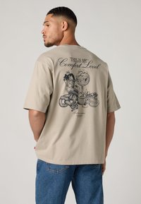 Smėlio spalvos medvilniniai marškinėliai su grafiniu atspaudu nugaroje, kuriame užrašyta "THIS IS MY Comfort Level" ir pavaizduoti desertai. Trumpomis rankovėmis.