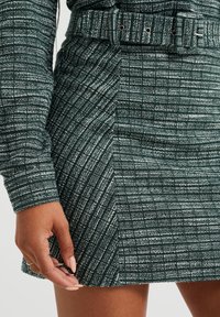 Minifalda a cuadros verdes y blancos con una tela texturizada, que presenta una cintura con cinturón y un detalle de abertura lateral. Mano posicionada en el dobladillo.