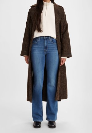 Femme portant un long manteau marron, un pull blanc à demi-fermeture éclair, un jean évasé bleu et des chaussures noires, debout devant un fond uni.