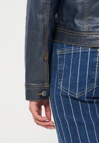 Mørk denimjakke med gul søm, med knappeerm. Kombinert med blå jeans prydet med vertikale rader av små, blanke nagler.