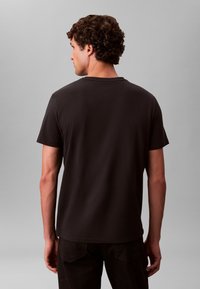 Mustast puuvillast t-särk lühikeste varrukatega, ümar kaelusega ja pehme tekstuuriga, vaadatuna tagant, kombineeritud tumedate pükstega.