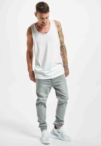 Top sin mangas blanco, pantalones ajustados grises claros y zapatillas blancas con detalles en negro. Presenta tatuajes en los brazos y está en una pose de pie.