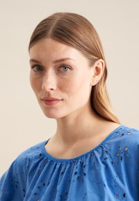 Blå bomuldsbluse med broderede mønstre og udskårne detaljer. Den har en rund halsudskæring og falder løst over skuldrene.