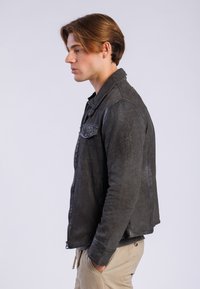 Chaqueta de cuero negro con una textura suave, cremallera frontal y cuello con solapa. Presenta un bolsillo en el pecho y un diseño ajustado.